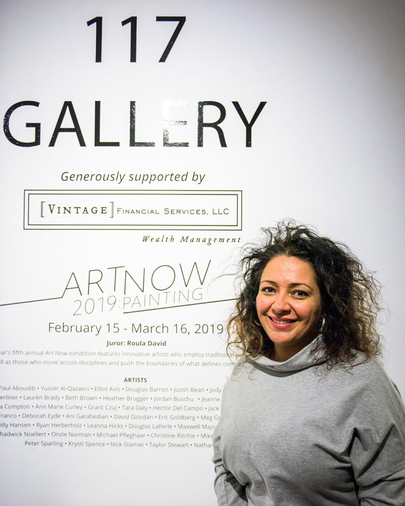 Juror Roula David standing next to exhibition title wall