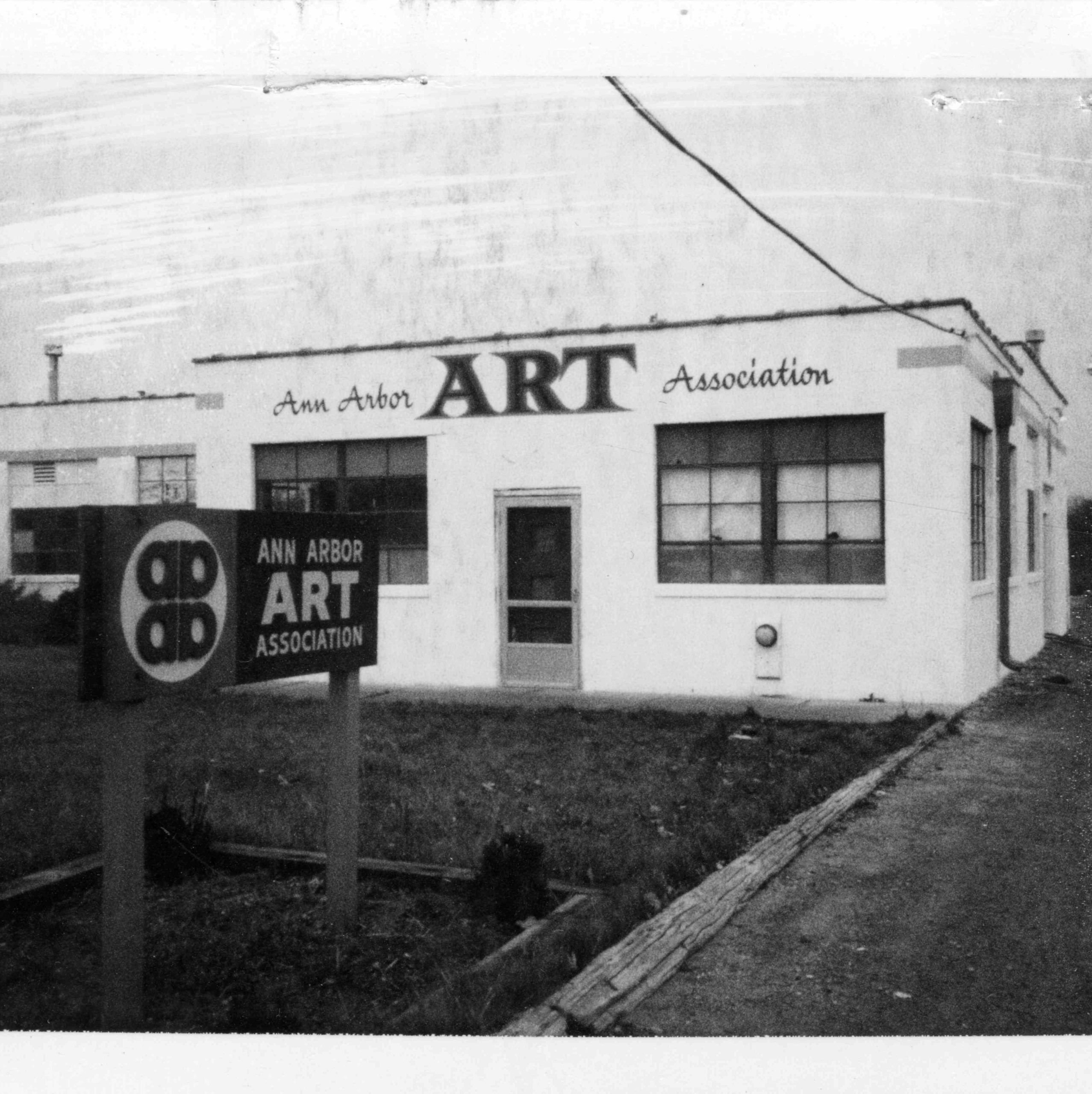 A2AC Historical Image. Black and White photo with a small white building in the middle of the photo with a sign and name on the building that says Ann Arbor Art Association (name before A2AC).