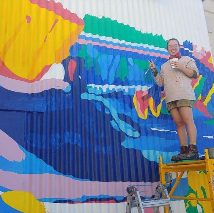Photo of Emily Herr painting a mural. She is standing on a yellow scaffolding with a brush on one hand, a cup on the other and she is making a peace sign with her right hand. The wall she is painting shows various colors predominantly blue, green, yellow, white and pink. The painting is abstract with organic, flowing, and curvy shapes.