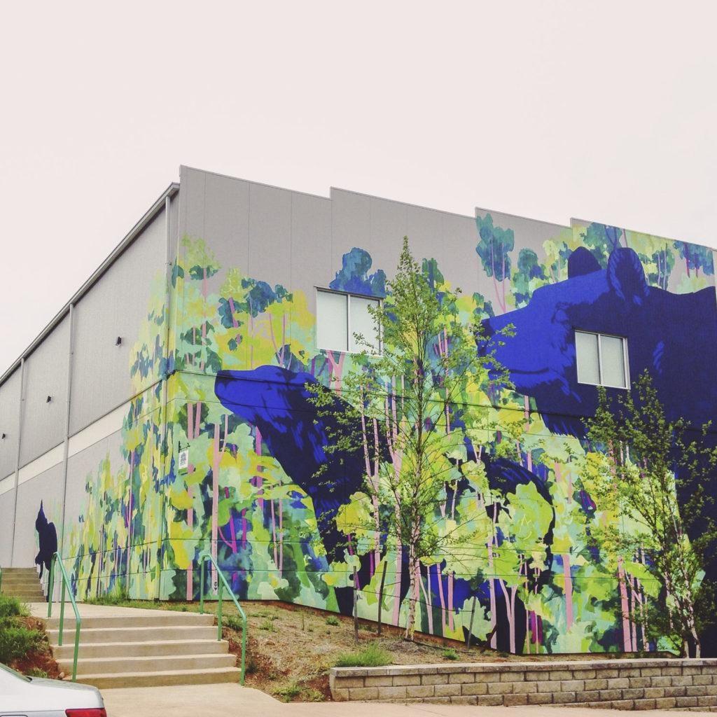 Photo of a mural for "TomTom Fest 2017." The mural is on a corner of a large warehouse covering an entire surface of the wall, curving around the corner and extending onto the other wall. The painting contains a series of green trees with pink barks and three blue bears strolling through the trees (one giant bear on the right of the wall, one smaller bear on the middle, and one smallest  bear at the left of the wall.