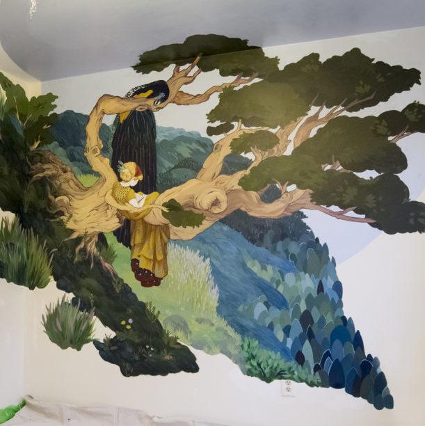 Photo of "Fantasia Drive" mural. The background is filled with green and blue hills. In the foreground there's a large tree growing on the side of a cliff.  On this tree there's a little girl with white skin and blonde hair wearing long golden dress while reading a book and a peacock looking at her read from a higher branch on the tree. 