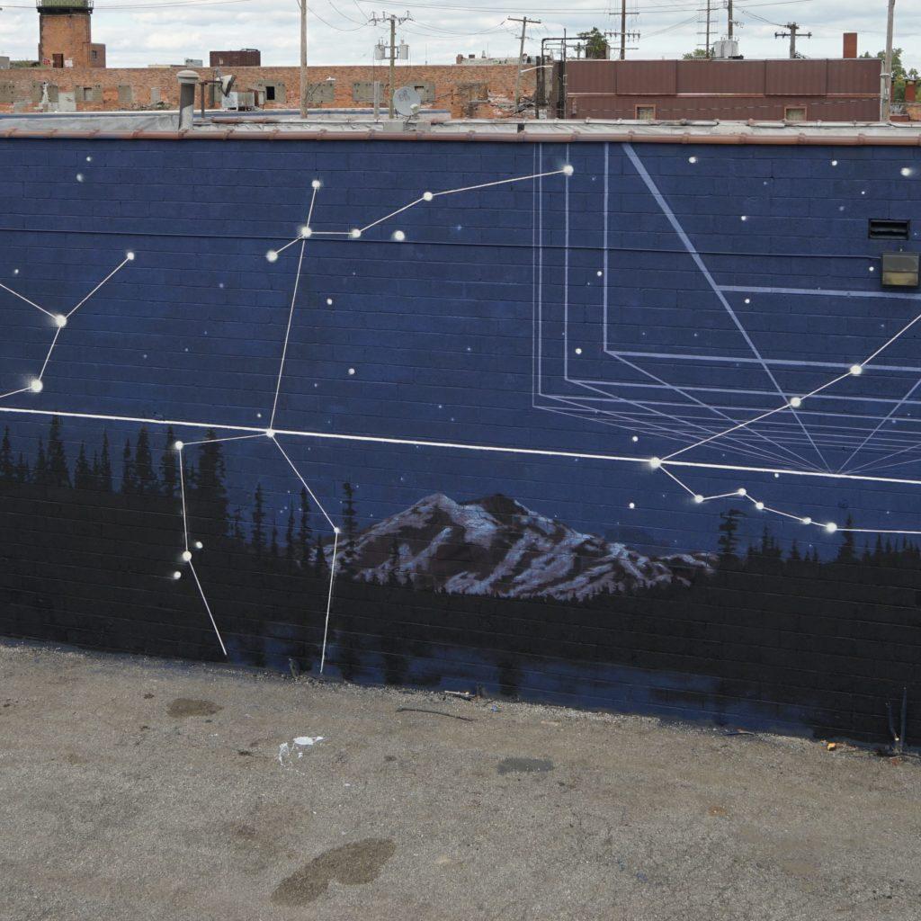 Photo of "Murals in the Market" mural. This mural shows a night time image of Picture Lake, with a reflection of Mount Shuksan in the calm water surrounded by pine trees.  The sky is dark with large stars connected with glowing lines. These stars and lines make up various constellation formations. 