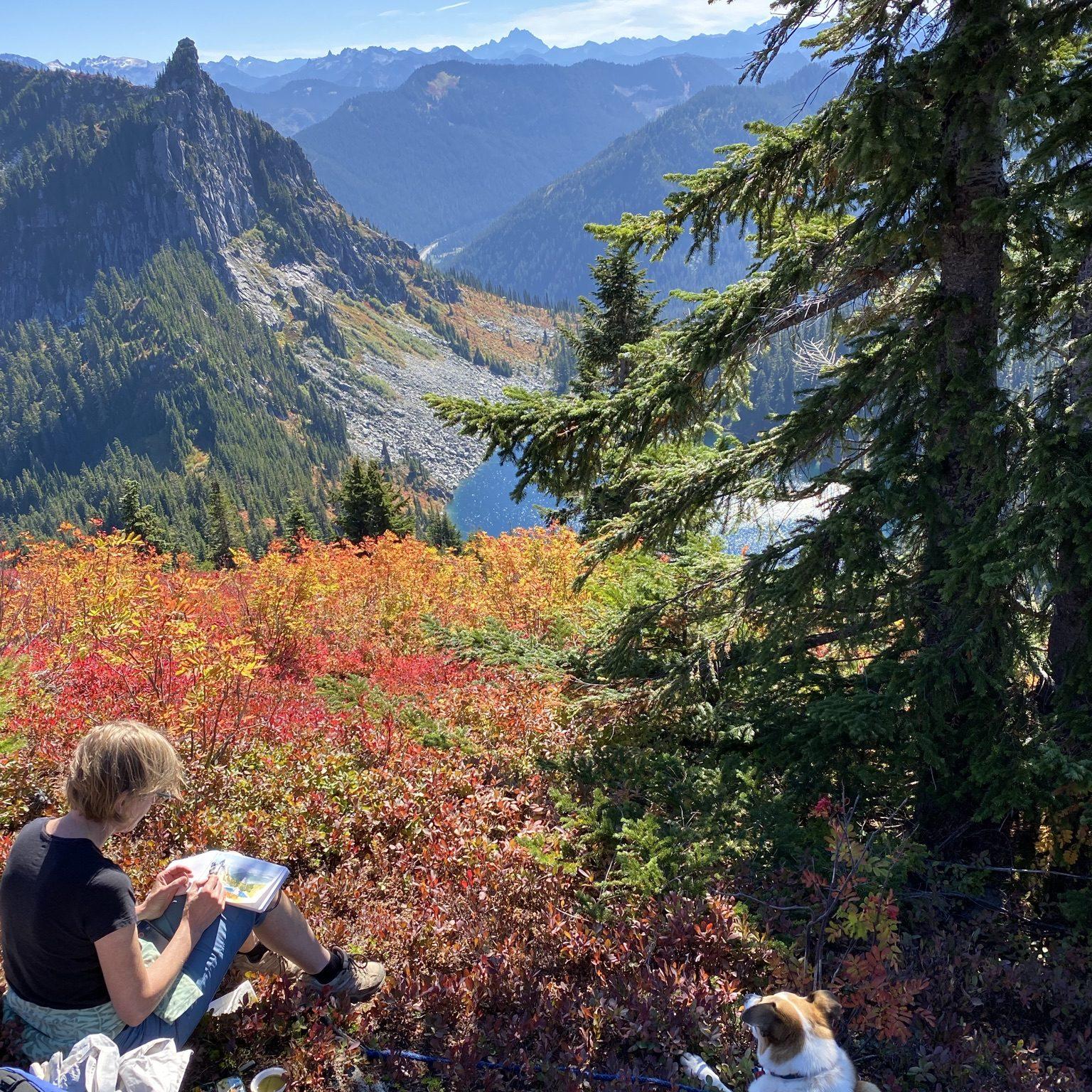 Photo of Mary Iverson and her dog on top of a hill looking our at mountains, pine trees, and a large river flowing through the mountains.